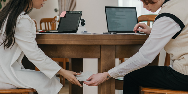 Two people sitting at a table with laptops. They are passing a note under the table.