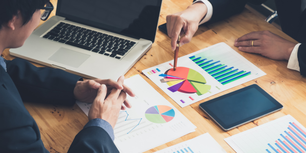 Two people are sitting at a table with papers with charts and graphs, a tablet and a laptop. One person uses a pen to point to a pie chart.