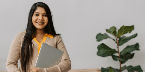 Person smiling and holding a laptop. There is a green plant on the right.