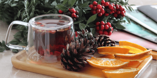 A clear glass mug filled with tea on a wooden tray, surrounded by pine cones, greenery, and berries, with the RIBO logo displayed in the foreground.