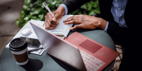 Person dressed in professional attire holding a pen and writing in a notebook. There is a laptop, coffee cup and other items on a table.